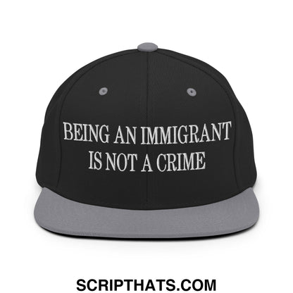 Black snapback hat with gray brim featuring white embroidered text being an immigrant is not a crime