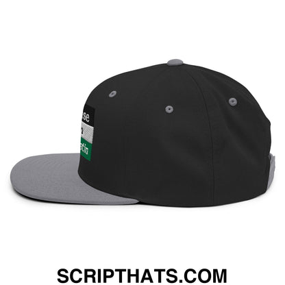 Black snapback cap with gray brim and rectangular patch with white text over green and black stripes on the side