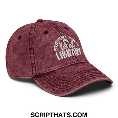 Maroon distressed denim dad hat with white embroidered support your local library text and a small book icon