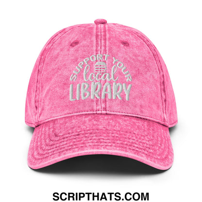 A faded pink denim dad hat with white embroidered support your local library text and a small library icon