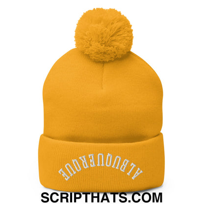 A mustard-yellow knit beanie with a fluffy pom-pom and white embroidered text on the cuff